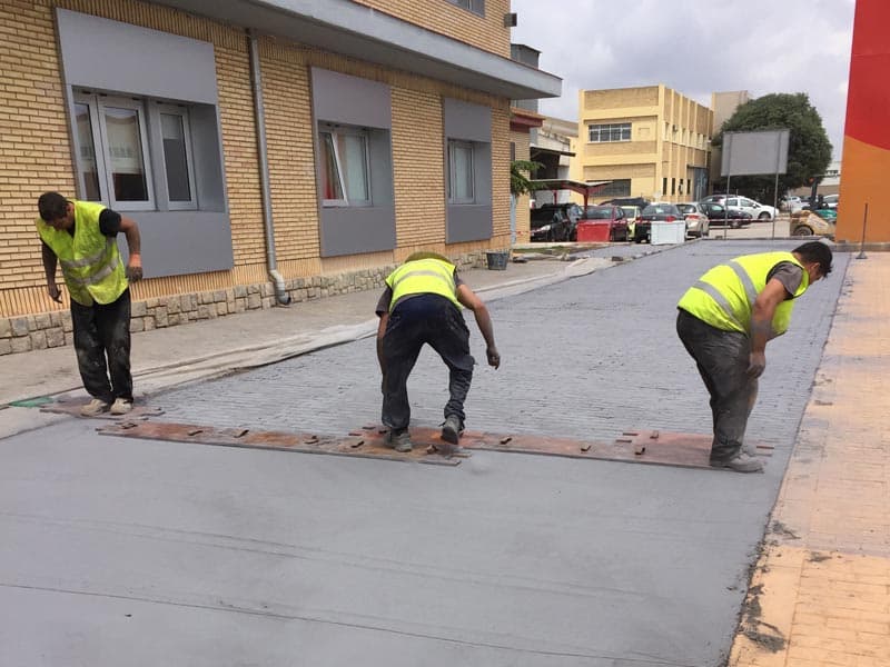 Tres operarios reglando a mano una solera de hormigón fresco en una acera, con el encofrado todavía puesto.