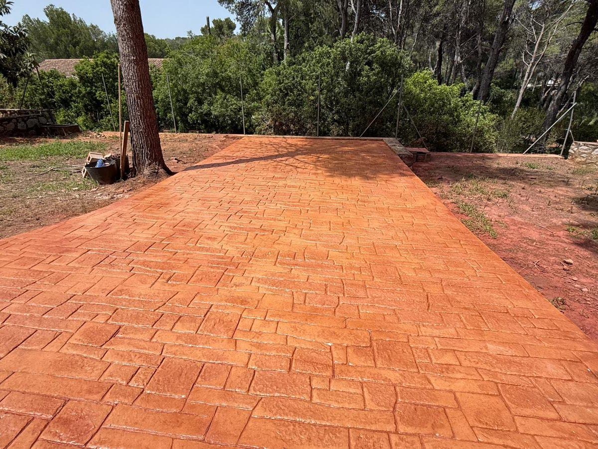 Explanada de hormigón impreso en adoquín irregular de tono rojizo, entre pinos.