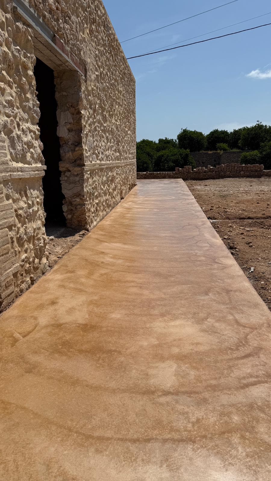 Paso de hormigón fratasado de tono arena junto a la fachada de piedra de una masía.