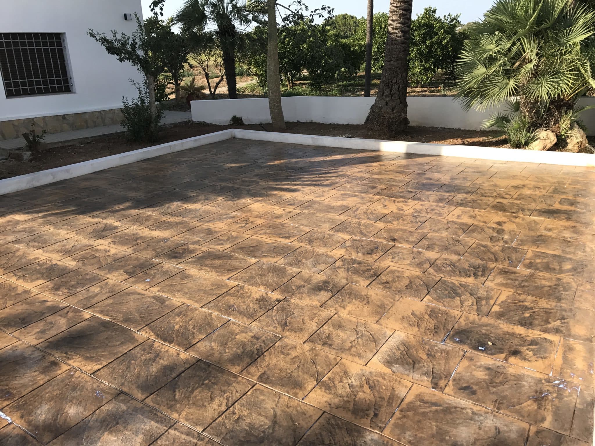 Patio de hormigón impreso en piedra inglesa de tono gris ante una vivienda blanca con palmeras.