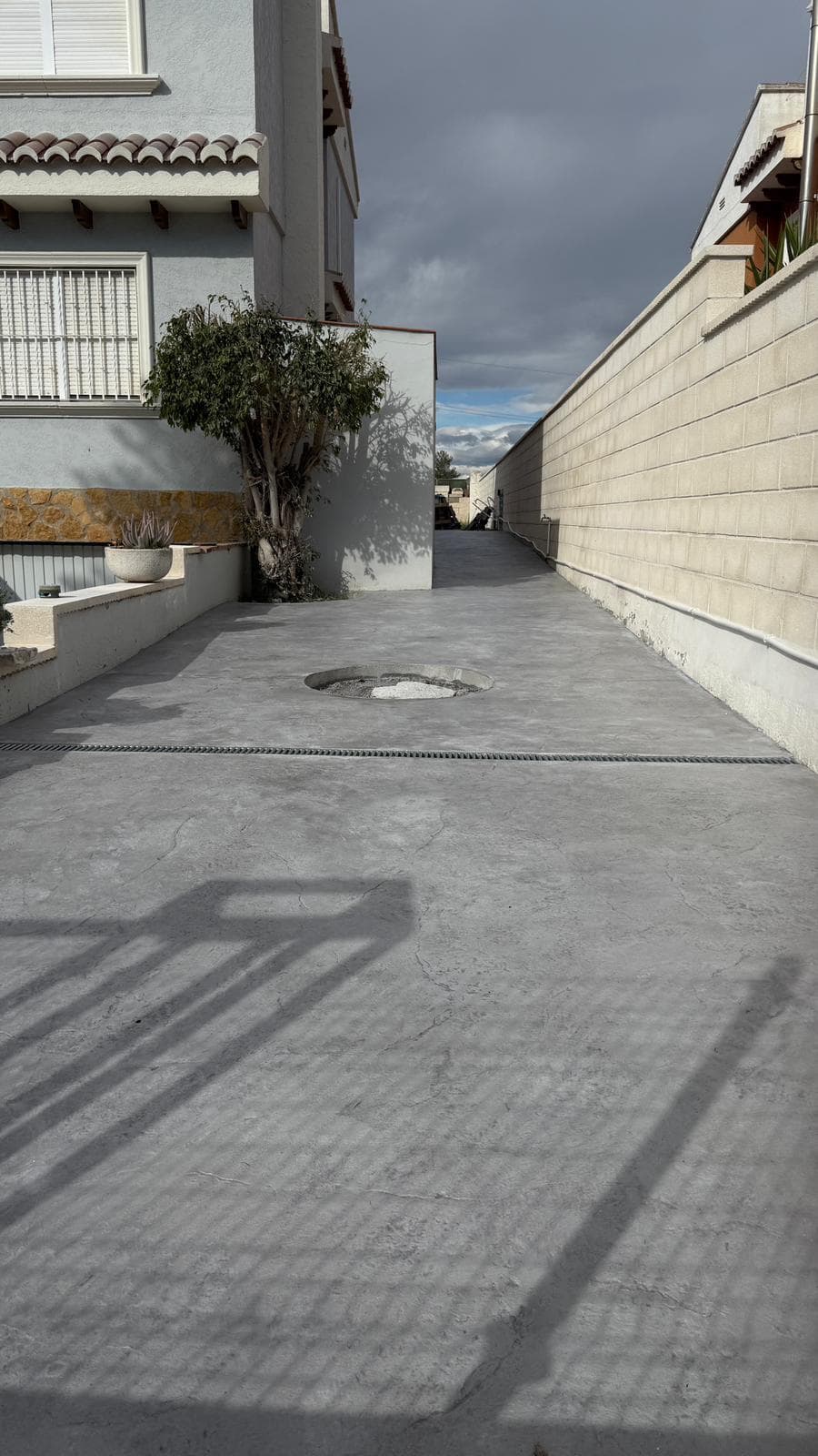 Paso lateral de hormigón gris continuo entre la fachada de una vivienda y el muro de la parcela.